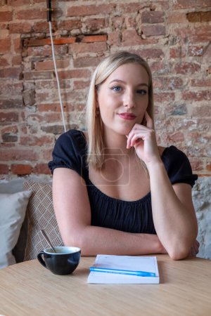 Téléchargez les photos : La photographie représente une jeune femme caucasienne aux cheveux blonds assise à une table dans un café, sa main touchant pensivement ses lèvres alors qu'elle regarde dans l'appareil photo. Une tasse de café noire se trouve à côté d'elle - en image libre de droit