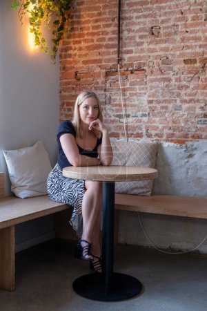 Cette image représente une jeune femme caucasienne aux cheveux blonds assise à une table dans un café chic, son regard réfléchi suggérant une pensée profonde ou contemplation. La lumière chaude et ambiante et la traînée