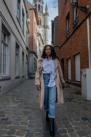 La imagen captura a una joven mujer negra caminando con confianza por un callejón empedrado en una zona urbana histórica. Ella está vestida a la moda con una camisa abotonada azul claro, pantalones vaqueros de ajuste relajado, y un largo