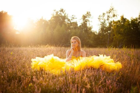 Eine kontemplative Frau sitzt inmitten eines Wildblumenfeldes, in das sanfte, goldene Licht der untergehenden Sonne getaucht, ihr leuchtend gelbes Kleid verschmilzt mit den goldenen Tönen der Natur. Goldene Träume