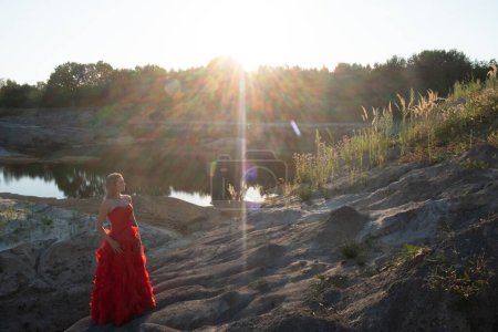Les rayons du soleil couchant projettent une lueur rayonnante sur une femme dans une robe rouge époustouflante, posée sur un terrain accidenté au bord du lac, résumant un moment d'élégance dans la nature. Elégance au milieu de la nature : Femme en rouge