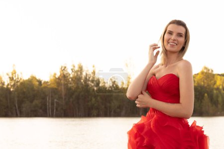 Eine Frau lächelt sanft und strahlt Anmut in einem leuchtend roten Kleid vor der weichen, natürlichen Palette eines ruhigen Seeufers in der Abenddämmerung aus. Lakeside Serenity: Anmutige Frau in einem fließenden roten Kleid. Hochwertiges Foto