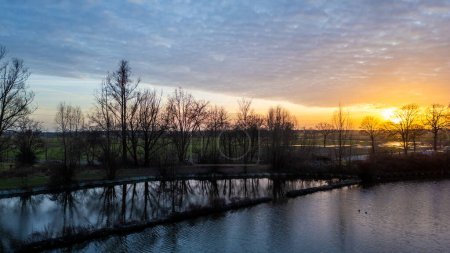 This image captures a serene dusk setting by a riverside, featuring the silhouettes of trees against a softly glowing sky. The sun, near the horizon, bathes the clouds in a gradient of orange and