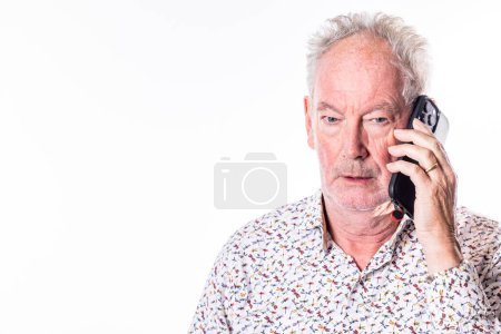 Un homme mûr semble engagé dans une conversation téléphonique sérieuse, son expression une contemplation concentrée, sur un fond blanc minimaliste. Homme âgé dans une conversation réfléchie au téléphone