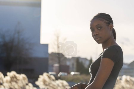 Das Foto zeigt eine nachdenkliche Afroamerikanerin bei Sonnenuntergang im Freien. Ihr geflochtenes Haar drapiert sich über ihre Schultern, und sie trägt ein dunkles Hemd, das mit dem frühen Abendlicht verschmilzt. Die Verschwommenheit