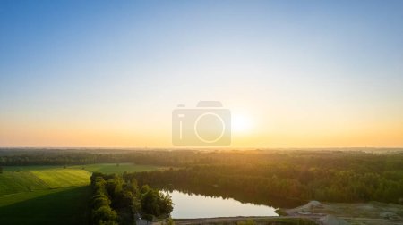 Cette photographie présente la première lumière de l'aube se brisant sur un paysage champêtre serein. Un petit lac se trouve tranquillement au milieu des champs et des arbres, reflétant le ciel tôt le matin. La douce lumière de