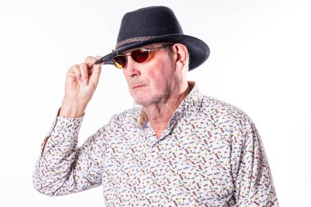 The image captures an elderly Caucasian man tipping his dark fedora hat with one hand. He wears sunglasses with a red tint, adding an air of mystery to his appearance. His attire includes a white