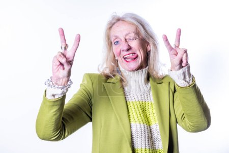 Portrait of an elderly woman radiating happiness, gesturing peace signs with both hands against a white backdrop, clad in a chic green jacket and knitted scarf. Joyful Senior Woman Making Peace Signs