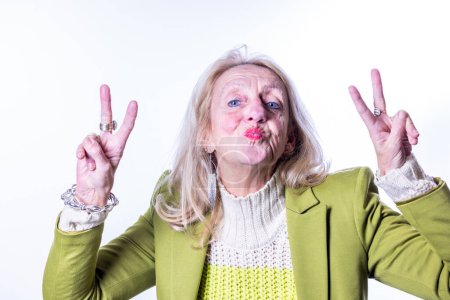 Une image attachante d'une femme blanche âgée avec de longs cheveux blonds donnant le signe de paix avec les deux mains. Elle caresse ses lèvres pour un baiser ludique et regarde directement la caméra avec une lumière