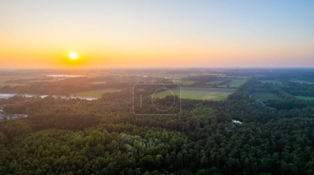This image captures the serene beauty of a sunset horizon viewed from above a verdant forest. The setting sun dips towards the horizon, its warm hues painting the sky in gradients of orange, yellow