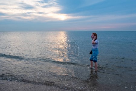 Esta imagen representa a un joven de pie en las aguas poco profundas del mar, con las manos suavemente colocadas sobre sus oídos, como si escuchara mejor el susurro de las olas. Están mirando hacia fuera
