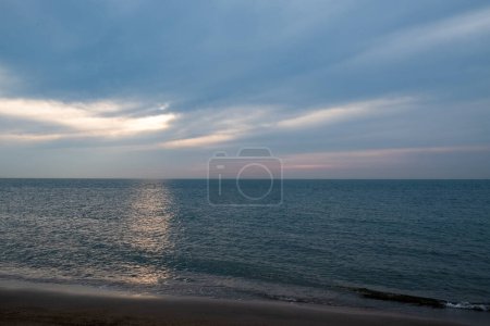 This image exudes tranquility through a serene seascape where the last rays of the sun pierce through parting clouds, casting a soft, ethereal glow on the water. The beach in the foreground is a