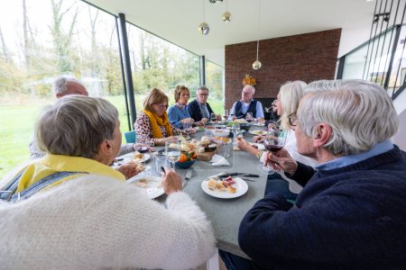 Group of adults engaged in conversation around a dining table, enjoying a meal together in a contemporary, well-lit space with a view of the outdoors. Family Gathering for a Meal in a Modern Sunlit