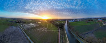 This panoramic aerial image captures a breathtaking sunset with a wide-angle view over a rural landscape divided by a serene canal. The setting sun bathes the scene in a soft golden light, contrasting