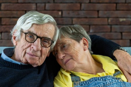 The image depicts an affectionate senior couple leaning on each other in a moment of relaxed intimacy. The man, with silver hair and glasses, shares the frame with a woman wearing a yellow top and