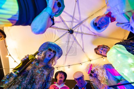 Una perspectiva única bajo la sombrilla captura un círculo de personas mayores con trajes vibrantes, cantando y disfrutando de una fiesta iluminada por luces de cuerda. Vista dinámica bajo el parasol de un disfraz de adulto mayor