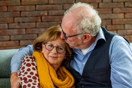 Ein liebenswertes Bild eines älteren Mannes in Weste und Brille, der seine Partnerin auf die Stirn küsst und herzlich lächelt. Die Frau mit der runden Brille und dem leuchtenden Senfschal zeigt eine