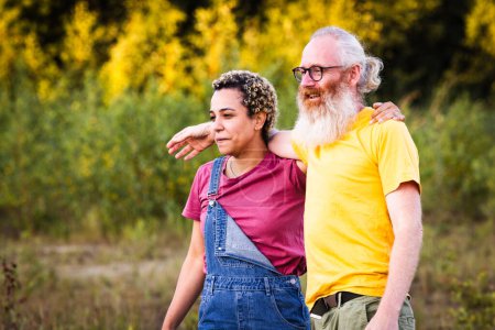 This image captures a caucasian man and a latina woman enjoying a peaceful moment in nature. The man, with a long white beard and glasses, stands behind the woman, who is wearing a denim overall dress