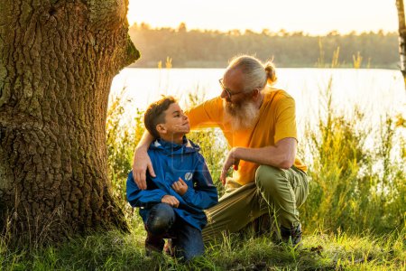 Dieses Bild fängt einen zarten Moment zwischen einem Großvater und seinem Enkel ein, der bei Sonnenuntergang an einem ruhigen Seeufer sitzt. Der ältere Mann mit weißem Bart, Brille und warmem Lächeln legt sanft seine Hand in den Mund.