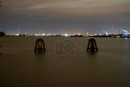 In this evocative night-time image, a panoramic view of a busy harbor is seen across calm waters. Industrial lights twinkle in the distance, creating a vibrant contrast against the dark evening sky. A