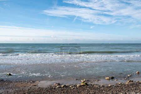 Dieses Foto fängt die Essenz eines friedlichen Tages am Strand ein und konzentriert sich auf eine Meereslandschaft, in der sanfte Wellen über eine mit Kieselsteinen beladene Küste spülen. Der Vordergrund ist übersät mit Steinen und Flecken von
