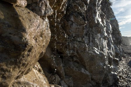 Esta imagen ofrece un detallado primer plano del agua que cae en cascada por una áspera y texturizada cara de roca en una playa soleada. La luz del sol atrapa gotitas individuales, destacando su claridad y los intrincados caminos