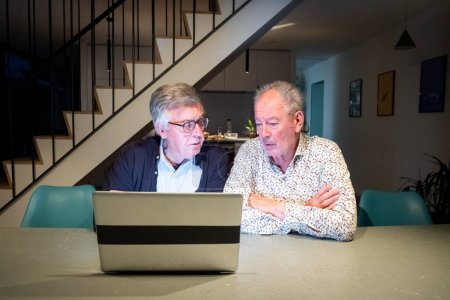 The image depicts two senior men focused on a laptop screen, possibly working together on a project or solving a problem. The man on the left, with glasses and a dark jacket, leans towards the screen