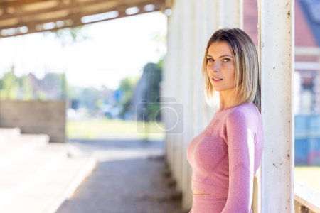 In the bright ambience of an urban space, a woman stands poised in form-fitting pink athletic attire, her focused expression capturing a moment of vigilance before activity. Urban Fitness Vigilance