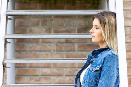 Profile view of a pensive young woman in a classic denim jacket, leaning near a window with architectural brickwork, embodying a moment of urban reflection. Urban Reflections: Casual Woman by the
