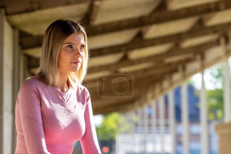 Una joven con un top rosa de manga larga y polainas se encuentra en un corredor urbano sombreado, su atención capturada por un punto distante, reflejando un momento de calma antes de la acción. Momento Reflexivo: Mujer