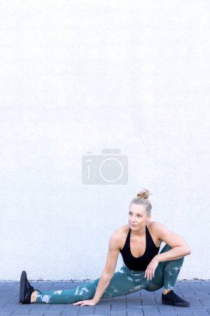 Eine schöne junge blonde Fitness-Frau in schwarz-blauer Sportkleidung führt eine Aufwärmübung durch, die tief auf den Boden fällt, um ihren Quadrizeps und ihre Schleimhäute ins Visier zu nehmen, was ihr morgendliches Training verbessert.
