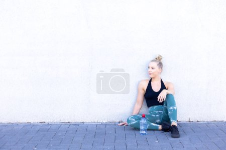 A happy and sporty young blonde millennial woman sits on the floor with a bottle of water after exercising, radiating wellness and a commitment to a healthy lifestyle while looking at the camera