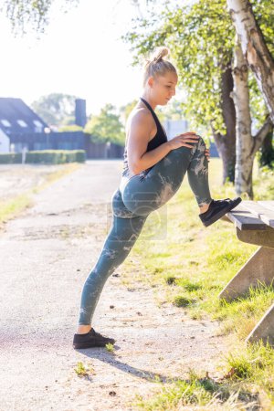 A young woman engages in a stretching routine on a sunny morning at a park. She places her foot on a bench, leaning forward for a deeper stretch, wearing a stylish activewear outfit. The natural