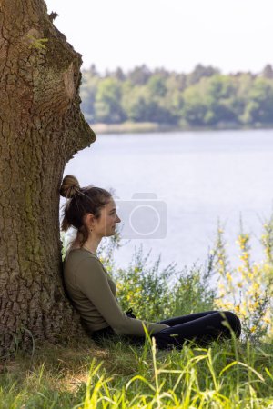 This image showcases a young woman in contemplation, sitting against a tree. The vastness of the lake and surrounding foliage creates a peaceful and introspective atmosphere. Contemplative Woman