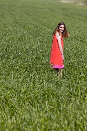 A smiling young woman stands in a field of tall green grass. She wears a bright coral dress, bringing a pop of color to the natural setting. Her relaxed posture and the expanse of the field evoke a