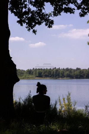 La imagen presenta una silueta llamativa de una mujer sentada en contemplación bajo un árbol, con la vista expansiva de un lago que se extiende ante ella. Las hojas crean un marco natural, encapsulando una