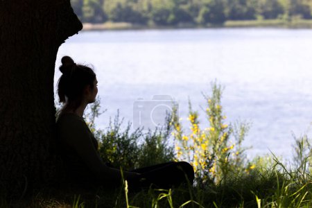 Captured in silhouette, a young woman sits serenely by a shimmering lake, gazing across the water as the day fades. The delicate balance of shadow and light accentuates the tranquility of the scene