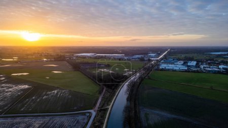 This is an inspiring aerial image showcasing a sunset over a serene landscape with a canal cutting through the farmland. The sun is low on the horizon, casting a warm glow and long shadows across the