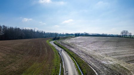The image presents an aerial view of a curvy, narrow road meandering through expansive fields. The fields appear dormant with remnants of snow or frost, suggesting this photo was taken during colder
