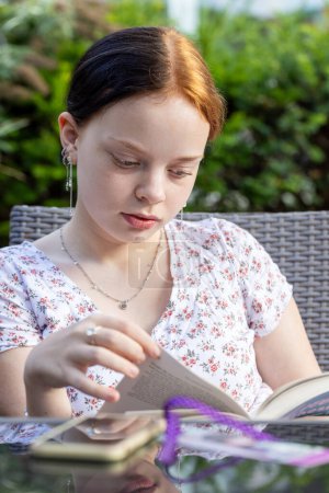 This intimate portrait captures a young redhead woman deeply absorbed in reading a book. Seated comfortably outdoors, her focused expression and the gentle turn of a page suggest a moment of quiet