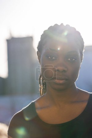 Una mujer africana está de pie retroiluminada por el sol poniente, su silueta parcialmente delineada contra el suave resplandor del cielo nocturno. Los rayos solares se filtran a través de su cabello, creando un efecto halo natural
