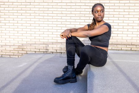 Eine afroamerikanische Frau sitzt selbstbewusst auf dem Boden, den Rücken vor einer weißen Backsteinwand. Sie trägt ein lässiges schwarzes Outfit gepaart mit Kampfstiefeln, das einen urbanen Straßenstil verkörpert. Ihre Zöpfe