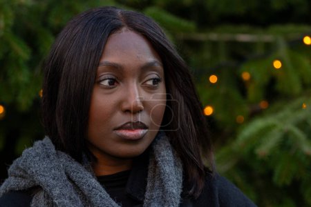 Captured against a backdrop of a fir tree adorned with twinkling lights, this image features a thoughtful African American woman in a warm black coat and a cozy gray scarf. Her gaze is directed off