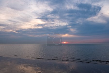 The photograph showcases a serene sunset descending over a calm seashore. The sun, a fiery orb, partially hidden by the cloud cover, casts a subtle reflection on the wet sand and gentle waves