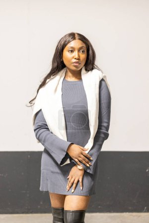 This image portrays a young woman with long, dark hair and medium-toned skin standing against a neutral background. She is wearing a form-fitting, grey knee-length dress with a white scarf draped over