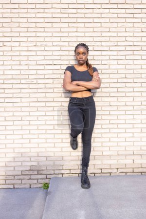 A young African woman stands before a brick wall, her feet firmly planted and arms crossed in a pose that exudes confidence and resilience. She wears an all-black ensemble comprising a close-fitting