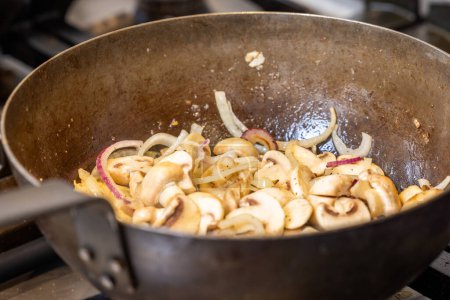 The image captures a close-up view of freshly sliced mushrooms and onions as they sizzle in a well-seasoned pan, which hints at many previous uses and flavorful meals. The steam rises subtly
