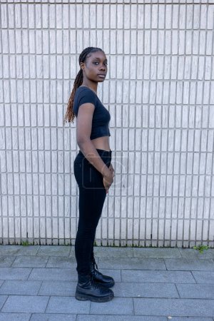 Téléchargez les photos : Cette image capture une jeune femme africaine debout avec confiance contre un mur de briques de couleur claire. Sa posture est détendue mais affirmée, car elle regarde directement dans la caméra avec une expression neutre - en image libre de droit