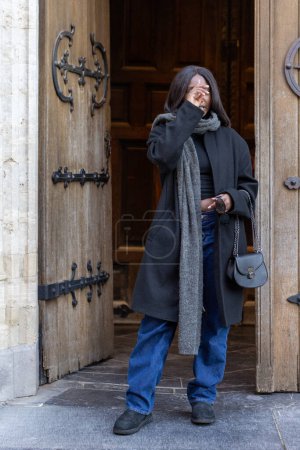 The image portrays a candid moment as a young woman, clad in a chic black coat and denim, steps out from the ancient doorway of a cathedral. With one hand delicately touching her face, she appears to