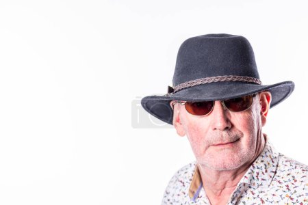 Portrait d'un homme âgé qui respire la confiance et le style, portant un chapeau de fedora et des lunettes de soleil sur un fond blanc vif. Les sujets sourire subtil et comportement détendu sont complétés par son